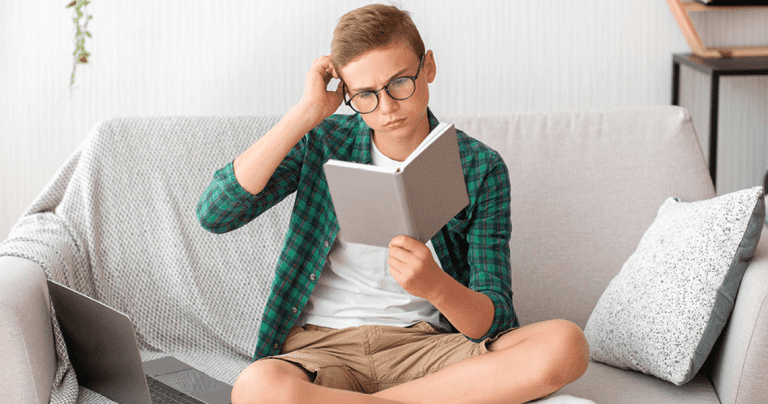 Boy reading book with laptop beside him